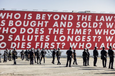 A group of soldiers beneath a red mural with text that begins 'WHO IS BEYOND THE LAW?'