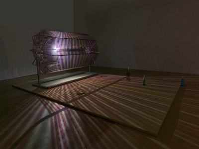 A sculpture composed of celluloid strips wrapped around a turning mechanism in a darkened room. A light pointed at the mechanism causes streams of purple light to shoot across the room, illuminating a bottle and assorted other objects on the floor.