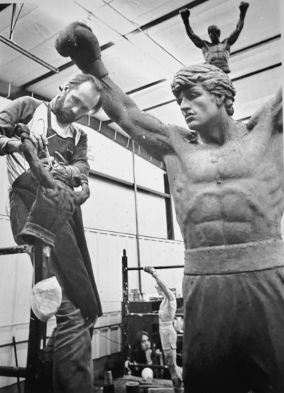 A photo shows bearded artist A. Gene Schomberg wearing an apron while working on a giant statue of Rocky Balboa.