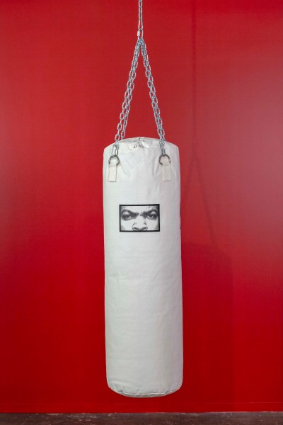 One photo shows a piece of art that includes a white sandbag with a black rectangle printed near the top depicting the eyes and nose of rapper and actor Ice Cooper. It hangs in front of a red background.