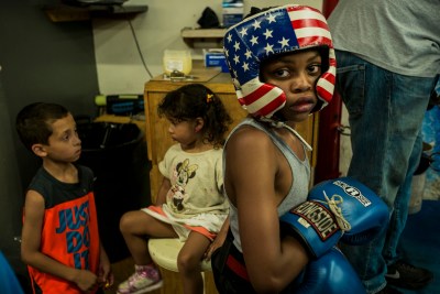 Three children pose for a photo at a boxing and martial arts studio in Philadelphia. One of the young black boys wears boxing gloves and a protective hat with stars and stripes