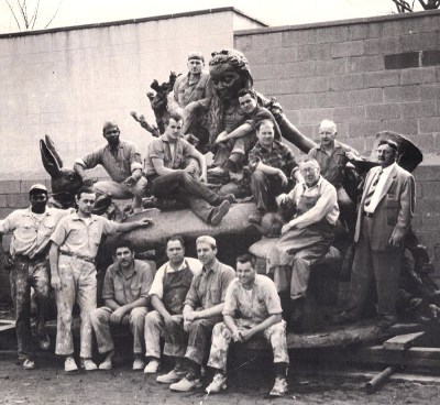 A group of male foundry workers sit atop an Alice in Wonderland sculpture. 