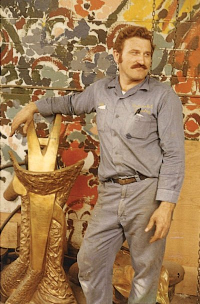 A man in workers clothing poses with gold-patinaed bronze sculptures. 