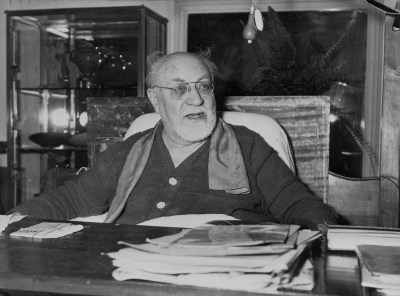 Portrait of Henri Matisse sitting at desk with books on it.