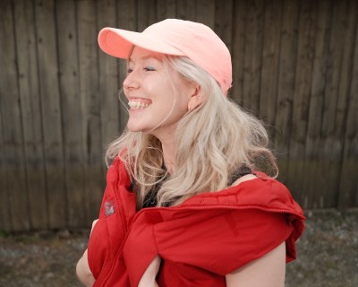 A smiling white woman in a pink baseball cap with her hands in her red vest's pockets.