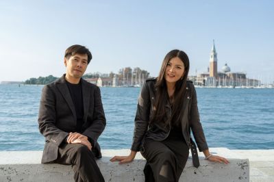 Two Hong Kong artists, Kingsley Ng and Angel Hui, dressed all in black, pose for the camera seated on a bench at the side of a canal in Venice, with the city's distinctive skyline in the distance