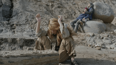 A film still showing a woman crouched down with her ams up in a desert landscape with two people behind her leaning on rocks.