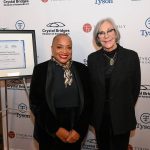 Two smiling women in front of a framed certificate.