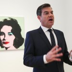 A man in a suit speaks in front of a painting.