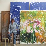 Artist Joan Mitchell, a white woman with brown hair wearing a denim shirt and jeans, poses in front of two large colorful abstract paintings in her studio