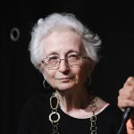 An older woman with white hair, glasses, black top and gold necklace