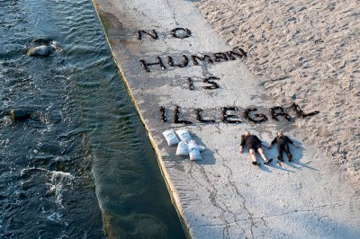 Two people lay on the banks of the Los Angeles River. Above them in ice soil reads 'NO HUMAN IS ILLEGAL'