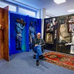 A photo shows artist Predrag Đaković sitting in his studio, wearing jeans, a leather jacket and sunglasses. Behind him are some works of art, mostly paintings with some three-dimensional elements, including figures, and some of playing musical instruments.
