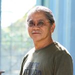 Portrait of a smiling indigenous man with a salt and pepper beard and round red-rimmed glasses. He was wearing a T-shirt and stood in front of the window, with light pouring in.