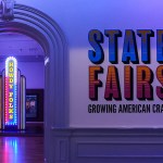 A neoclassical room under purple light has two signs: one on the wall reads "State Fair: Evolving American Crafts" Another light was on and read
