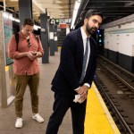 A man in a suit leans on the edge of the subway platform. A man behind him was checking his cell phone.