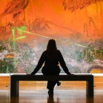 A woman sits on a bench in front of an orange landscape with lasers shining through it.