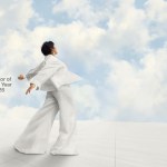 A man wearing a flowing white shirt and pants raises his arms backwards against an overcast sky.