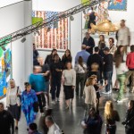 People walk through the aisles of a crowded art fair. Some people in the image have been blurred.