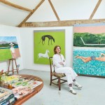 A woman dressed in white sits on a chair, surrounded by paintings.