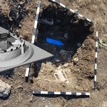 The photo shows an earth-covered ground with blue arrows pointing to archaeological remains and the reconstruction of the fortress