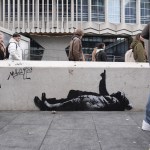 A piece of graffiti artwork depicting two children lying on the sidewalk in winter clothes, with one of them pointing to the sky, appears on a low wall in London in front of a Brutalist skyscraper. Numerous people are seen walking by.