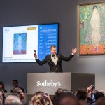 A man in a tuxedo raises his arms, a painting of a woman hangs behind him, and a screen to the left shows the current bids for the painting in multiple currencies.