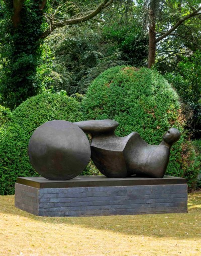 A photo shows Henry Moore's sculpture showing the stylized figure of a man lying on his back, made of black material, located in a sculpture garden