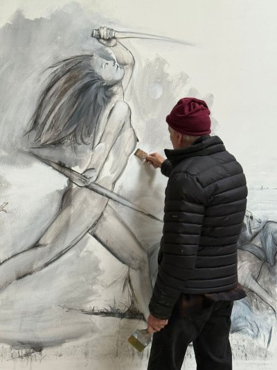 An artist applies gray paint to a large canvas depicting a naked woman holding a weapon.