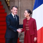 A smiling man in a dark blue suit and tie shakes hands with a smiling woman in a red dress and jacket, standing in front of a red staircase with flags in the background