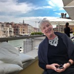 Curator Kathleen Goncharova sits by the canal on a sunny day in Venice, Italy, a white-haired caucasian woman smiles at the camera