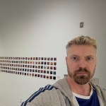 Nick Dwyer, a young white artist, poses in an art gallery with many small images pinned to the wall behind him