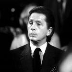 In the black and white photo, a man with dark hair, suit and tie looks sad