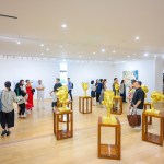 People walk around the gallery, where several golden sculptures sit on wooden plinths in the foreground.
