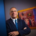 Portrait of John P. Axelrod, standing in a museum with a painting behind him (out of focus).