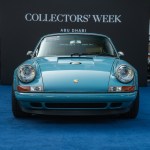 A light blue Porsche is parked on a dark blue carpet, surrounded by trees in the background.