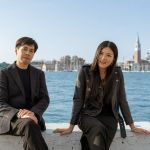 Two Hong Kong artists, Kingsley Ng and Angel Hui, dressed in black, pose for the camera while sitting on a bench by the canal in Venice, with the city's unique skyline in the distance