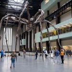 A large room has a sculpture of a giant spider and people walk underneath it.