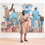 A black man dressed in completely nude clothing stands in front of a painting depicting various figures.