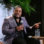 A black man speaks into a microphone next to a glass of water. He pointed with one finger. He wears a purple jacket with a diamond pattern on it.
