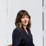 A woman in a black blazer sits and turns her head towards the camera against a white background.