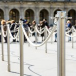 Stainless steel poles connected with gray ropes are used for queue control at the entrance of tourist attractions, with shadows on the light ground and people queuing blurred in the background.