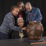 Four black men (three standing, one seated) examine 1850s ceramic vessels made by their ancestors.