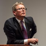 On the rostrum, a man in a suit leaned forward. He holds a pen in one hand.