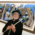 A man with a white beard stands in front of a painting.