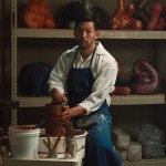 An Asian man wearing a dirty apron works at the pottery wheel. Abstract sculptures sit on metal shelves behind him.