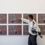 A man with short hair wearing dark pants and a white sweatshirt points to a row of framed photos hanging on a gallery wall