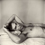 A vintage photo of a shirtless man lying on a bed, arms raised above his head, hairy armpits clearly visible.