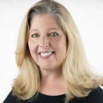 Mary Anne Carter, a blond Caucasian woman, smiles at the camera against a blank white background.