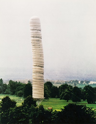 A tall white inflatable column towering above trees in a green field with a gray sky behind it